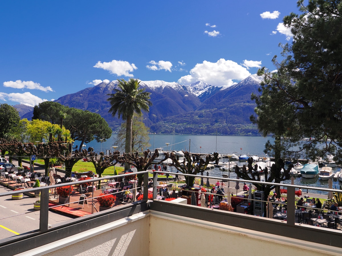 A scenic view of a lake is framed by majestic mountains capped with snow. Lush greenery can be seen below, along with a promenade populated by clustered chairs and tables under trees. The bright blue sky features scattered clouds, enhancing the tranquil setting.