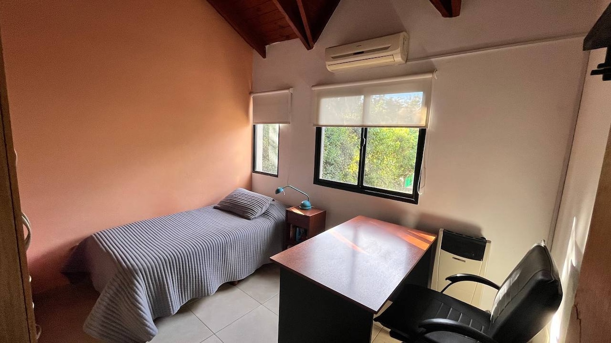 A well-lit bedroom features a single bed with a striped blanket. A desk with a comfortable black chair provides a workspace. Two windows allow natural light to enter, revealing views of greenery outside. An air conditioning unit is mounted on the wall.