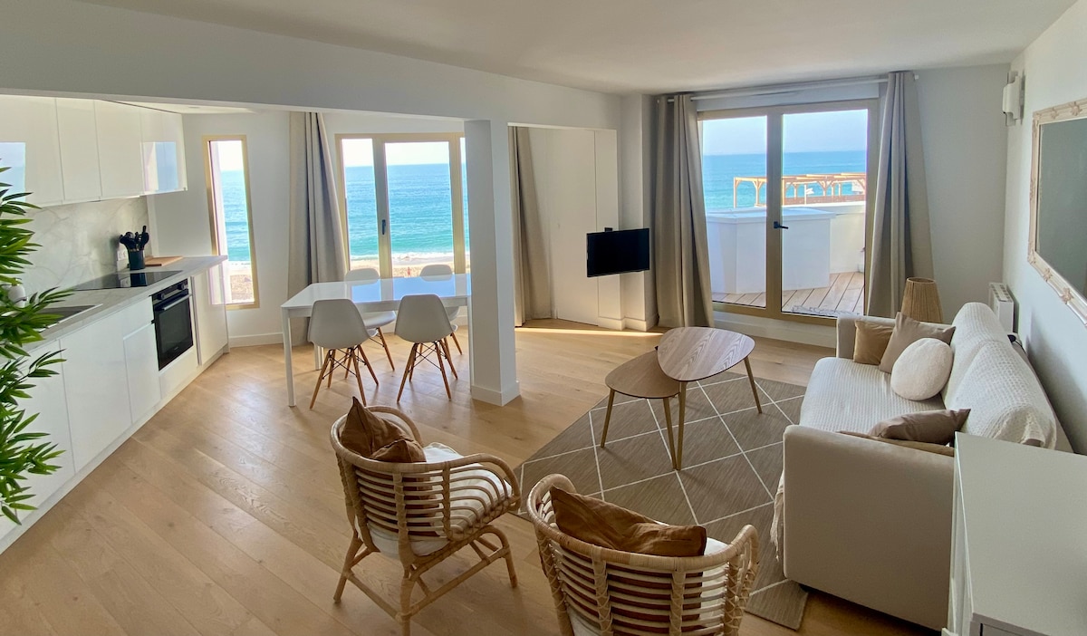 A spacious living area features large windows that provide a captivating view of the ocean. A modern kitchen with sleek cabinetry is complemented by a dining table for four. Two comfortable chairs and a soft sofa create a relaxed setting complemented by warm natural light.