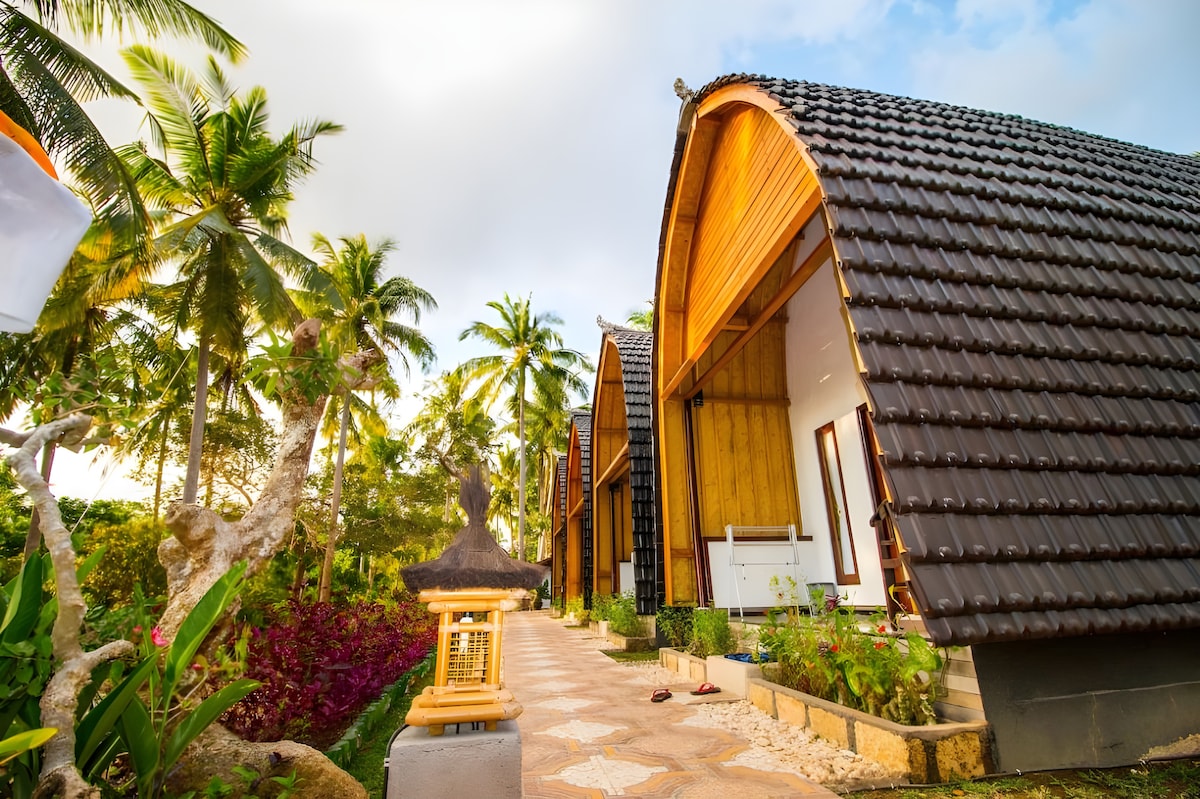Spacious bungalows are nestled among lush greenery and palm trees, featuring distinctive curved roofs. A stone pathway leads to the entrance, surrounded by vibrant plants and decorative elements. Soft sunlight filters through the clouds, enhancing the natural colors of the surroundings.