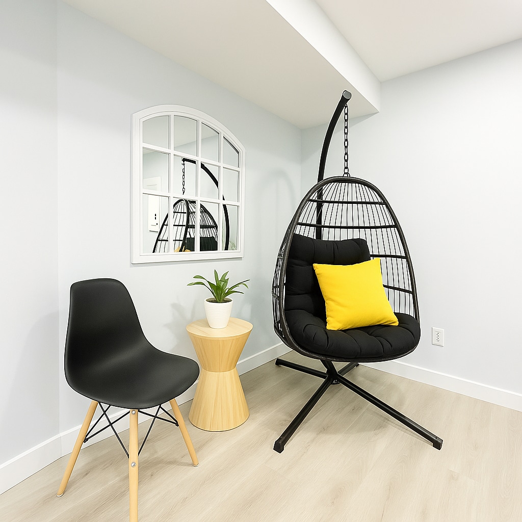 A relaxed seating area features a swing-style chair designed with a dark frame and soft black cushions. A bright yellow throw pillow adds a pop of color. Nearby, a small wooden table holds a potted plant, complementing the light grey walls and inviting atmosphere.