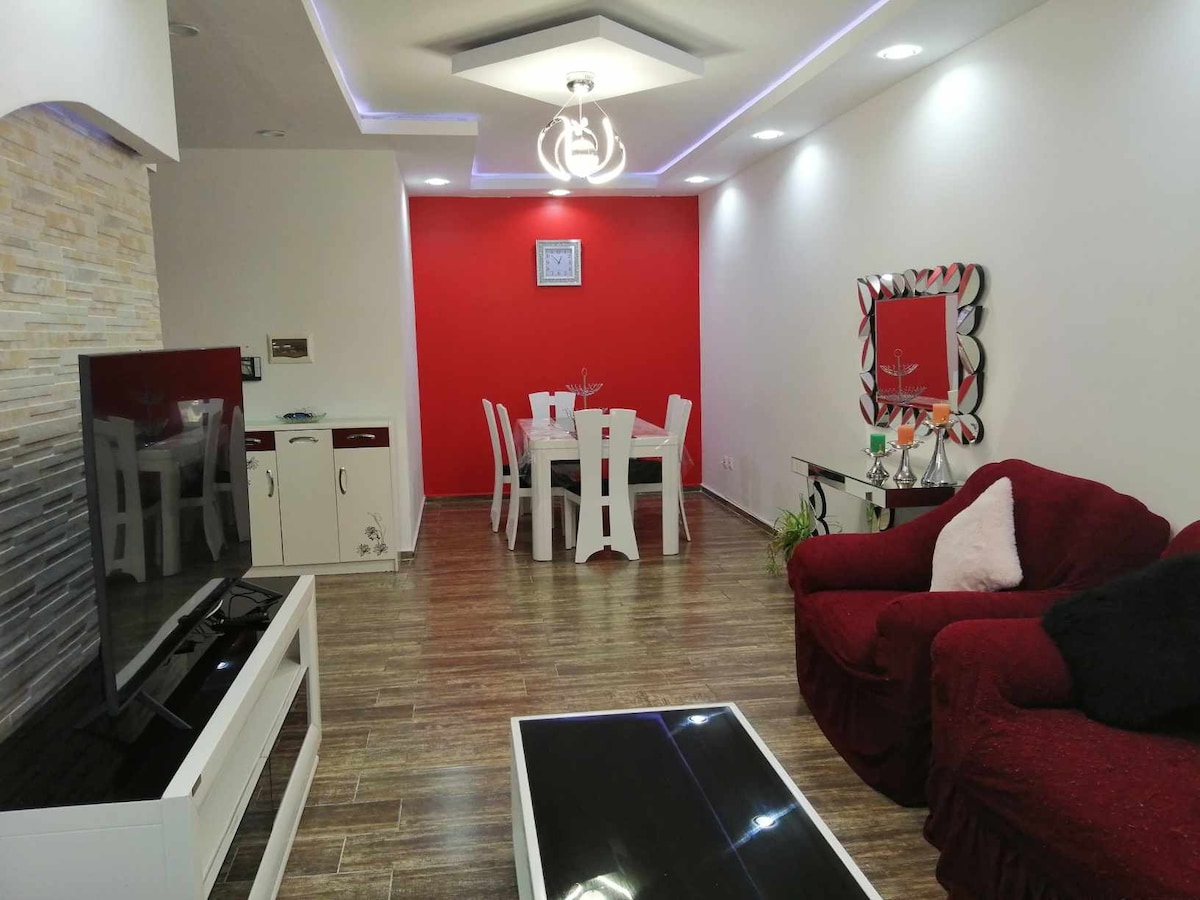 An inviting living and dining area features a red accent wall. A white dining table surrounded by chairs is positioned adjacent to a plush red sofa. Natural light is welcomed by the recessed ceiling and spotlighting, while a sleek black coffee table complements the modern decor.