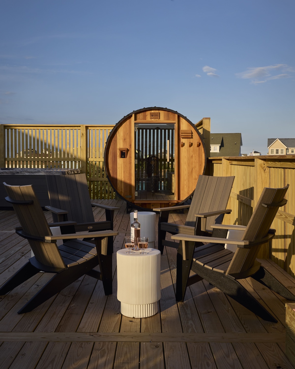 A spacious outdoor deck features two comfortable lounge chairs arranged around a small table. A circular sauna is prominently positioned in the background, set against a clear blue sky and surrounded by fencing that offers privacy.