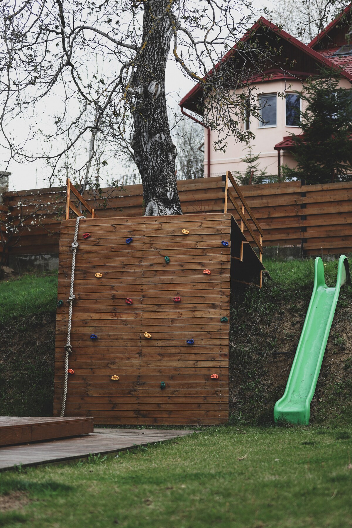 A wooden climbing wall features colorful handholds and a rope, situated adjacent to a green slide. The outdoor play area is bordered by grassy terrain, with a red-roofed house visible in the background, partially obscured by trees and a wooden fence.