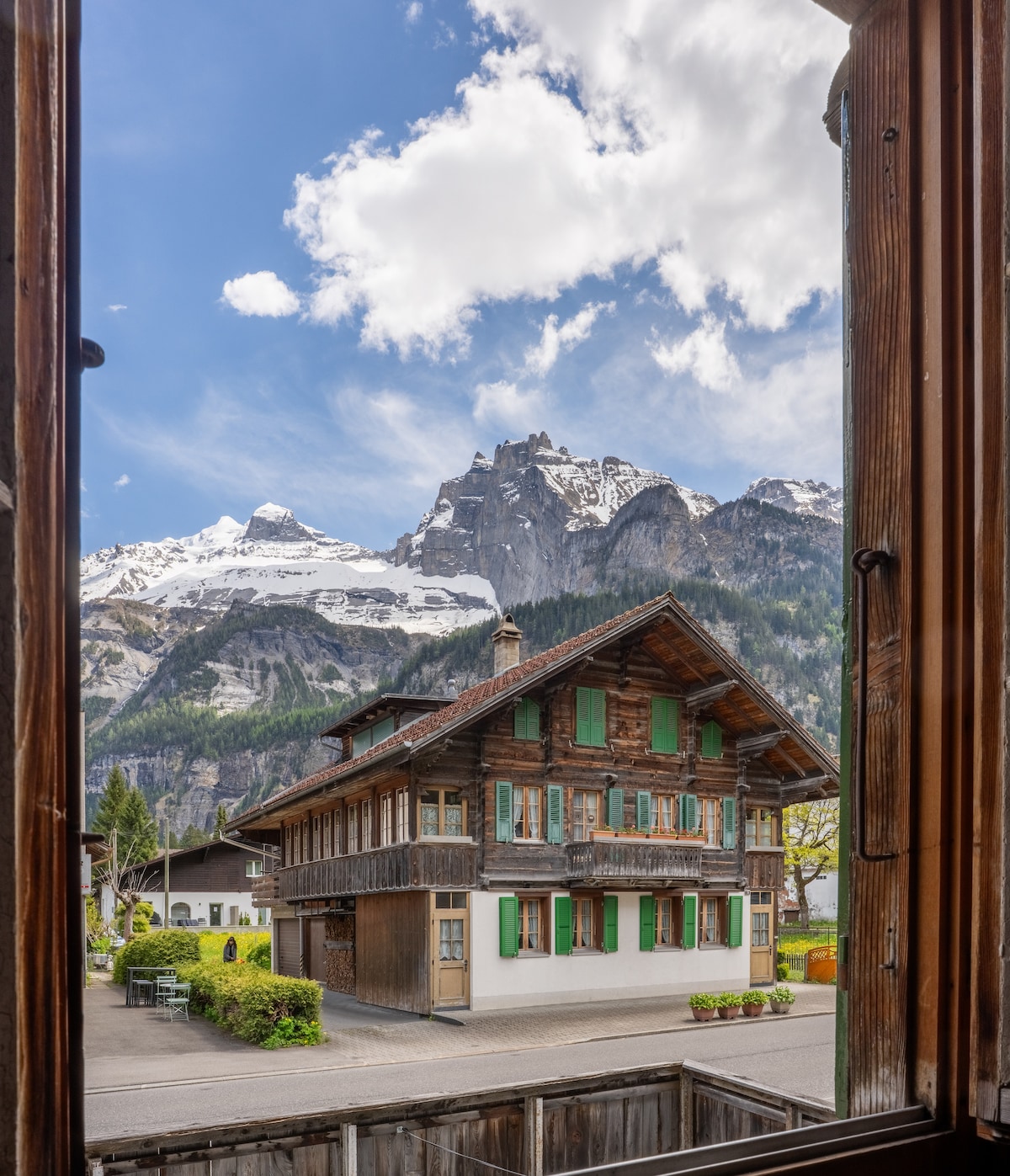 The view from a window frames a traditional wooden chalet with green shutters, surrounded by blossoming greenery. Majestic snow-capped mountains rise in the background under a partly cloudy sky, creating a serene alpine landscape.