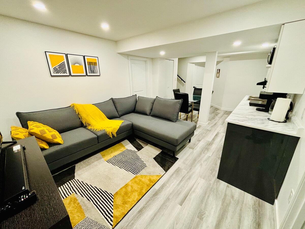 A cozy living area features a spacious L-shaped sofa adorned with a yellow throw blanket. A geometric area rug complements the modern decor, while a sleek kitchenette is visible in the background, equipped with essential appliances. Wall art in bold colors adds a pop of interest.