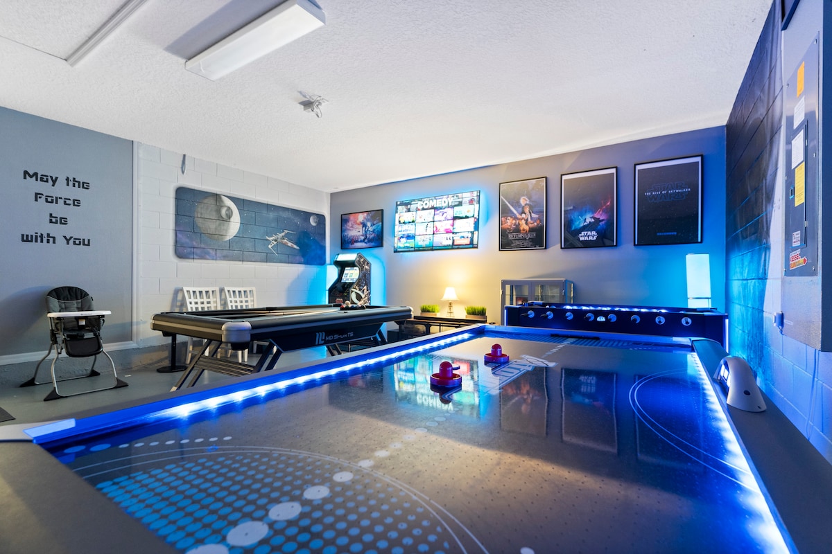 The game room features a modern air hockey table with illuminated edges, surrounded by a Star Wars arcade game and a treadmill. The walls are adorned with themed artwork, while a TV displays various games. Ambient lighting enhances the playful atmosphere.