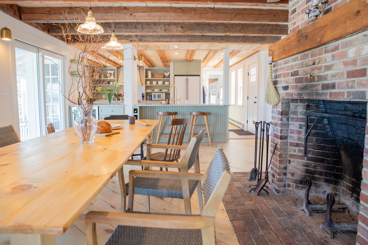 A spacious dining area features a large wooden table surrounded by comfortable chairs. The room is illuminated by hanging lights and boasts an open kitchen in the background. A rustic brick fireplace is situated to the side, complemented by a wooden beam ceiling.
