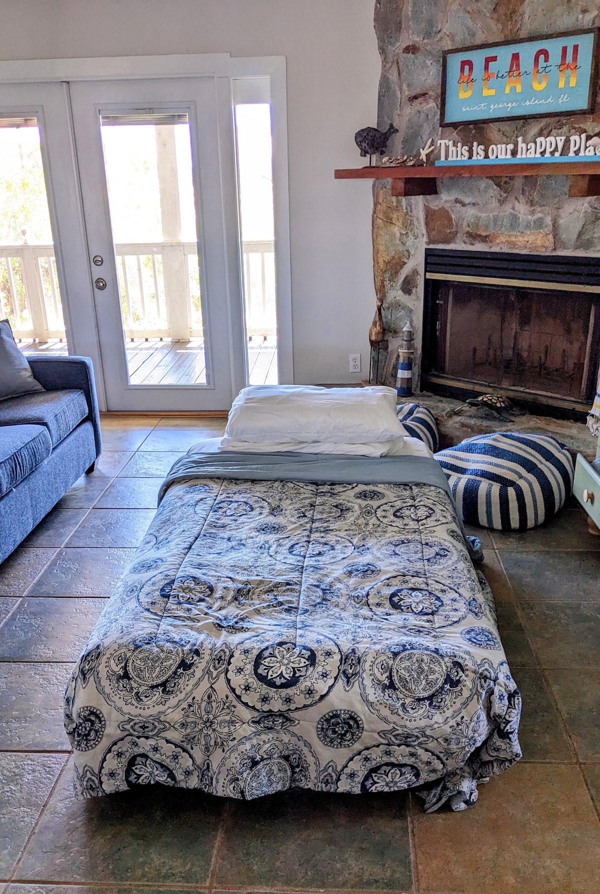 A spacious living area is highlighted by a comfortable sleeping space on the floor, featuring a patterned bedding set. A stone fireplace is visible along the back wall, and glass doors lead to a covered outdoor space. Decorative accents add character to the room.