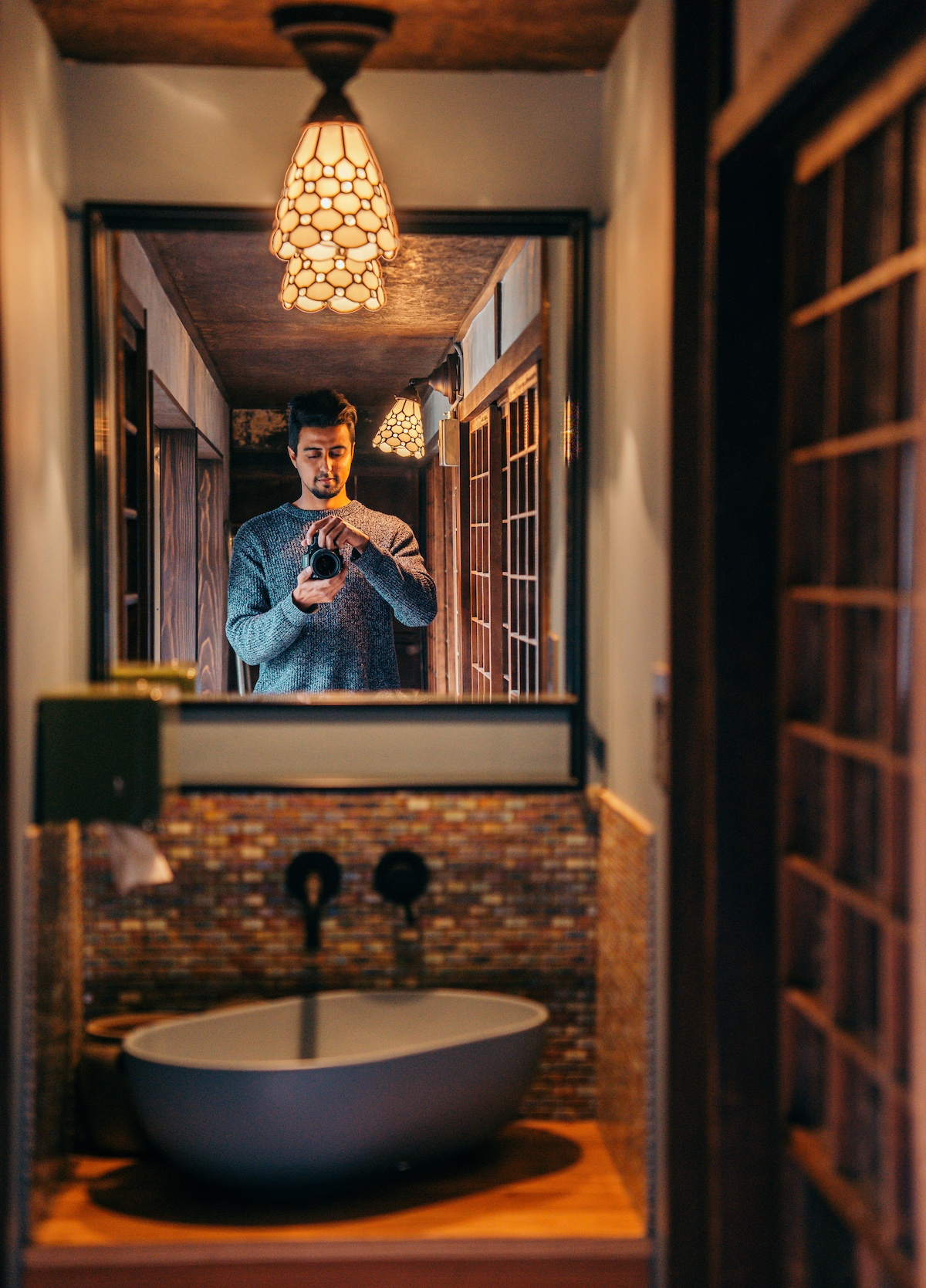 A modern bathroom is captured in the image, featuring a sleek, oval-shaped sink set upon a wooden countertop. Above, an intricately designed pendant light illuminates the space. A full-length mirror reflects the room's warm tones and wooden elements.