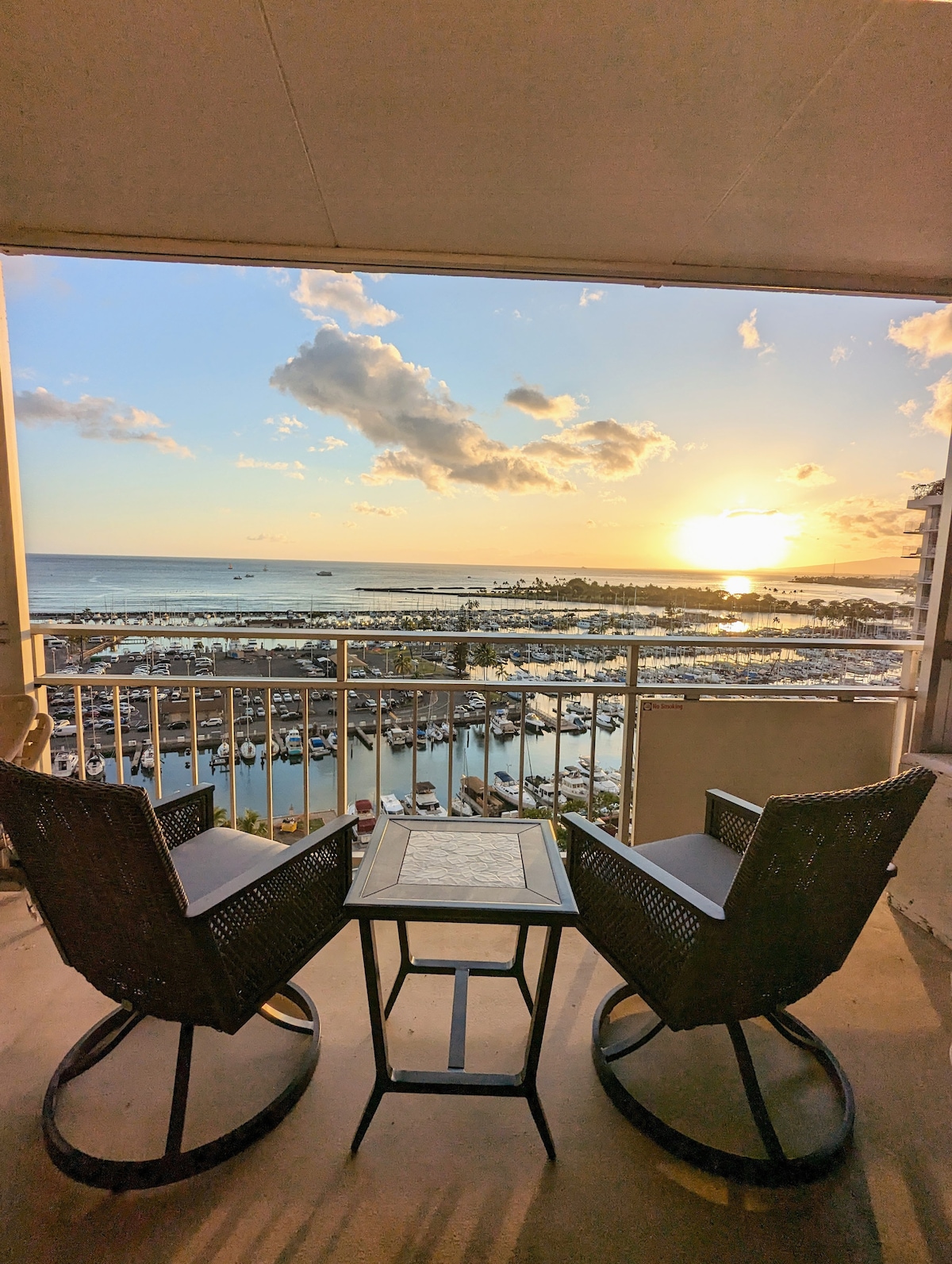 A lanai with two rocking chairs positioned beside a small table offers views of the marina and ocean at sunset. Soft light reflects off the water, creating a serene ambiance. The horizon is adorned with clouds, enhancing the scenic backdrop.