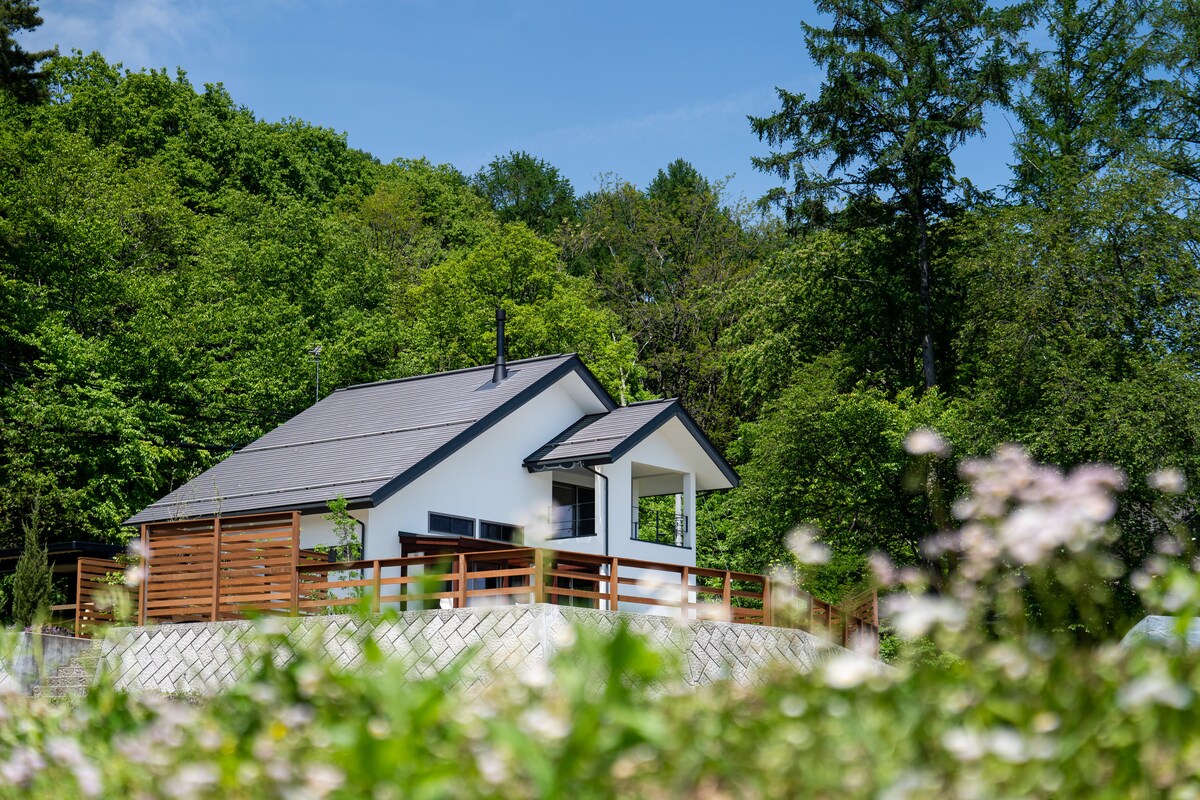 A modern, single-story villa is surrounded by lush greenery, featuring a welcoming porch with a sloped roof. The house displays a clean design with large windows, and a wooden deck complements its natural setting.