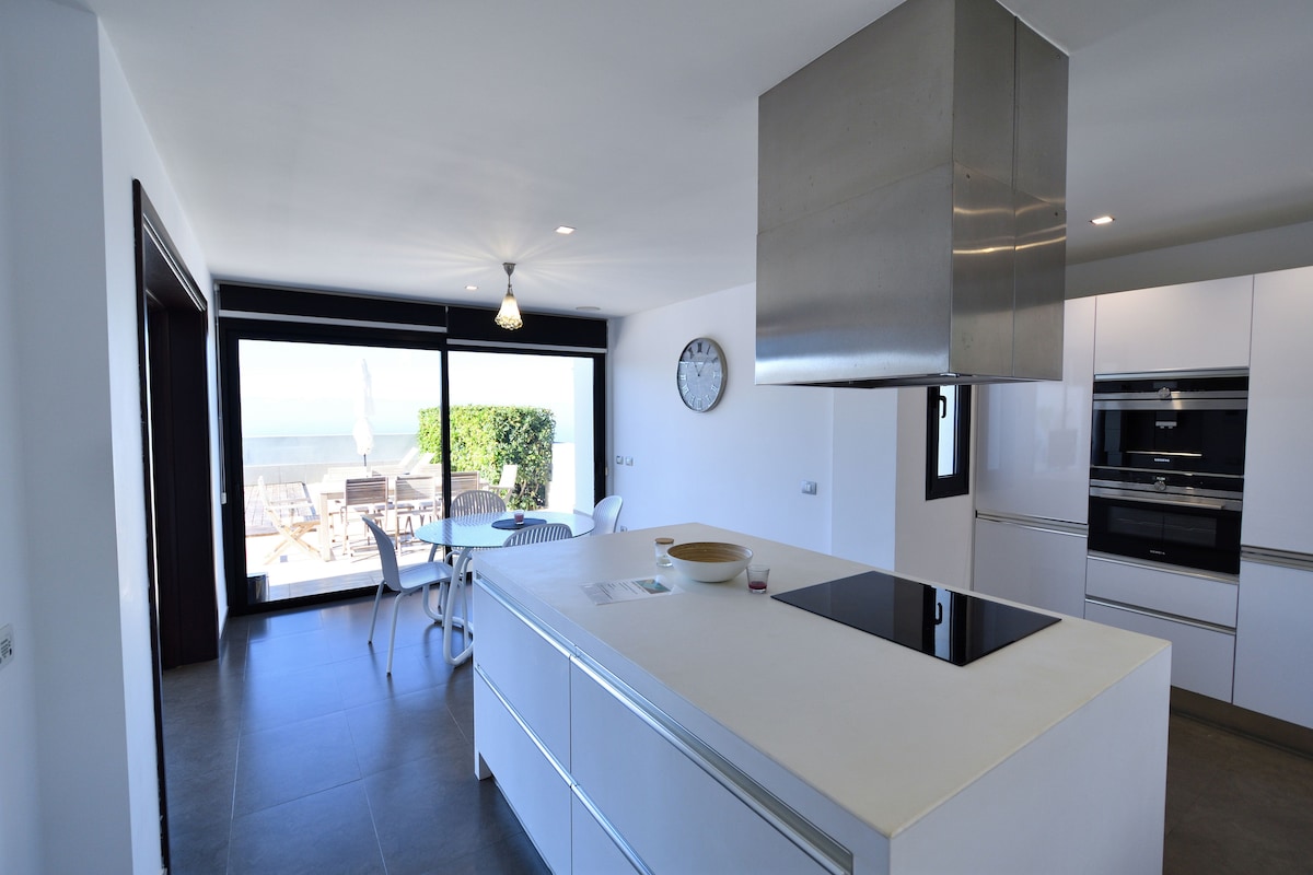 A modern kitchen features a central island with a cooktop, complemented by sleek cabinetry. Large floor-to-ceiling windows provide a view of the outdoor dining area and ocean. A wall clock is mounted above, adding to the contemporary design.