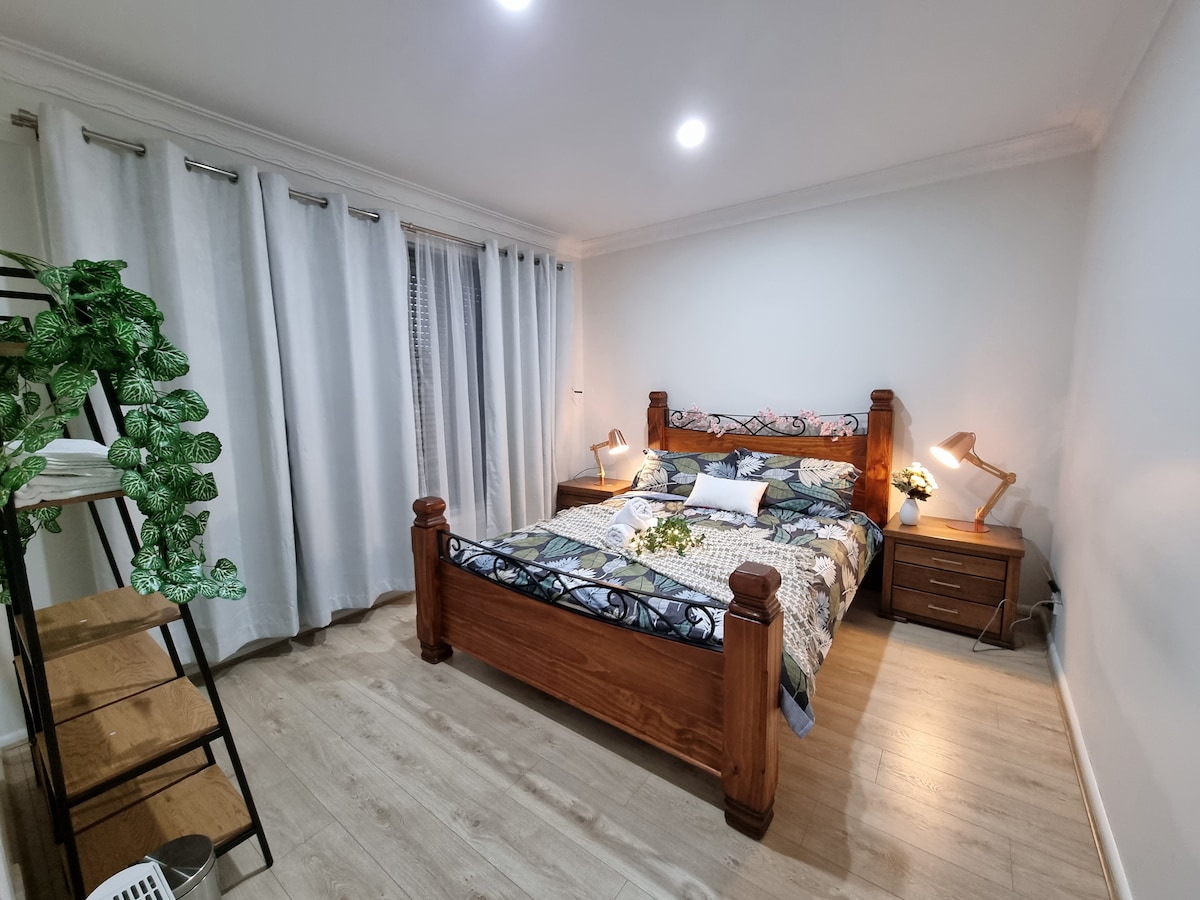 A well-appointed bedroom features a spacious bed framed in dark wood, complemented by decorative bedding. Natural light filters through sheer curtains, enhancing the serene ambiance. A bedside table with a lamp and decorative plants adds warm accents, while a modern shelving unit provides storage space.