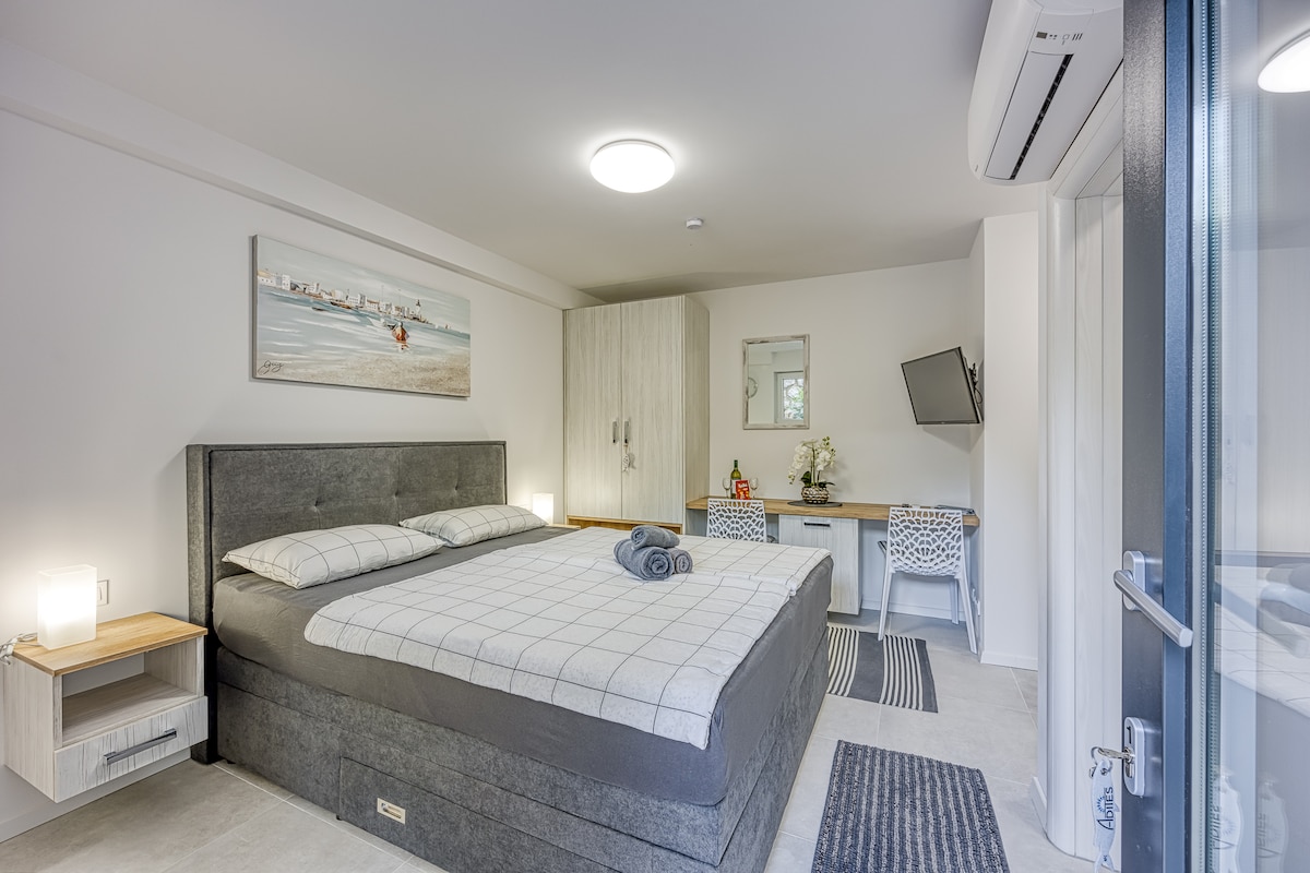 A spacious sleeping area features a large bed with a neutral-tone plaid bedding set. Two bedside lamps provide soft illumination, while a large painting depicting a serene beach scene adorns the wall. A small desk with two chairs is positioned next to a flat-screen TV.