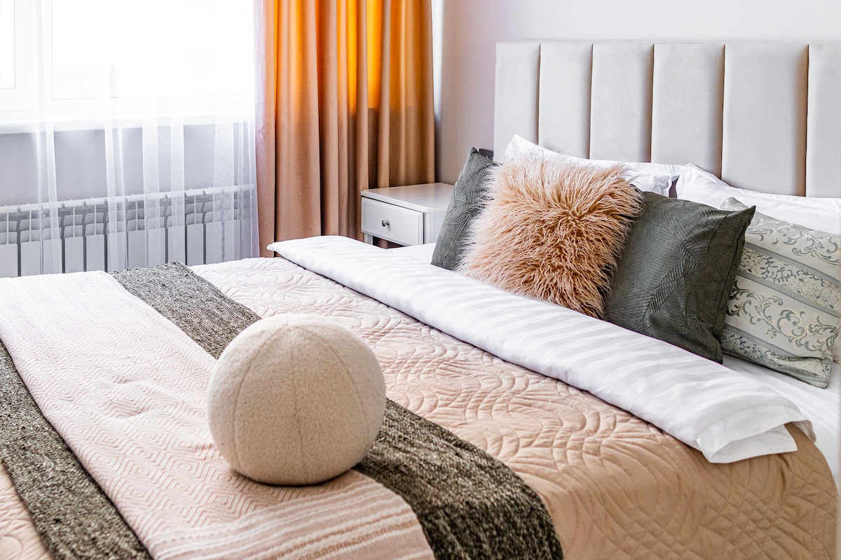 A cozy bedroom features a spacious bed adorned with soft, neutral-coloured linens and several decorative pillows. Natural sunlight enters through large windows draped with sheer curtains, creating a warm ambiance. A bedside table complements the decor, adding to the inviting space.