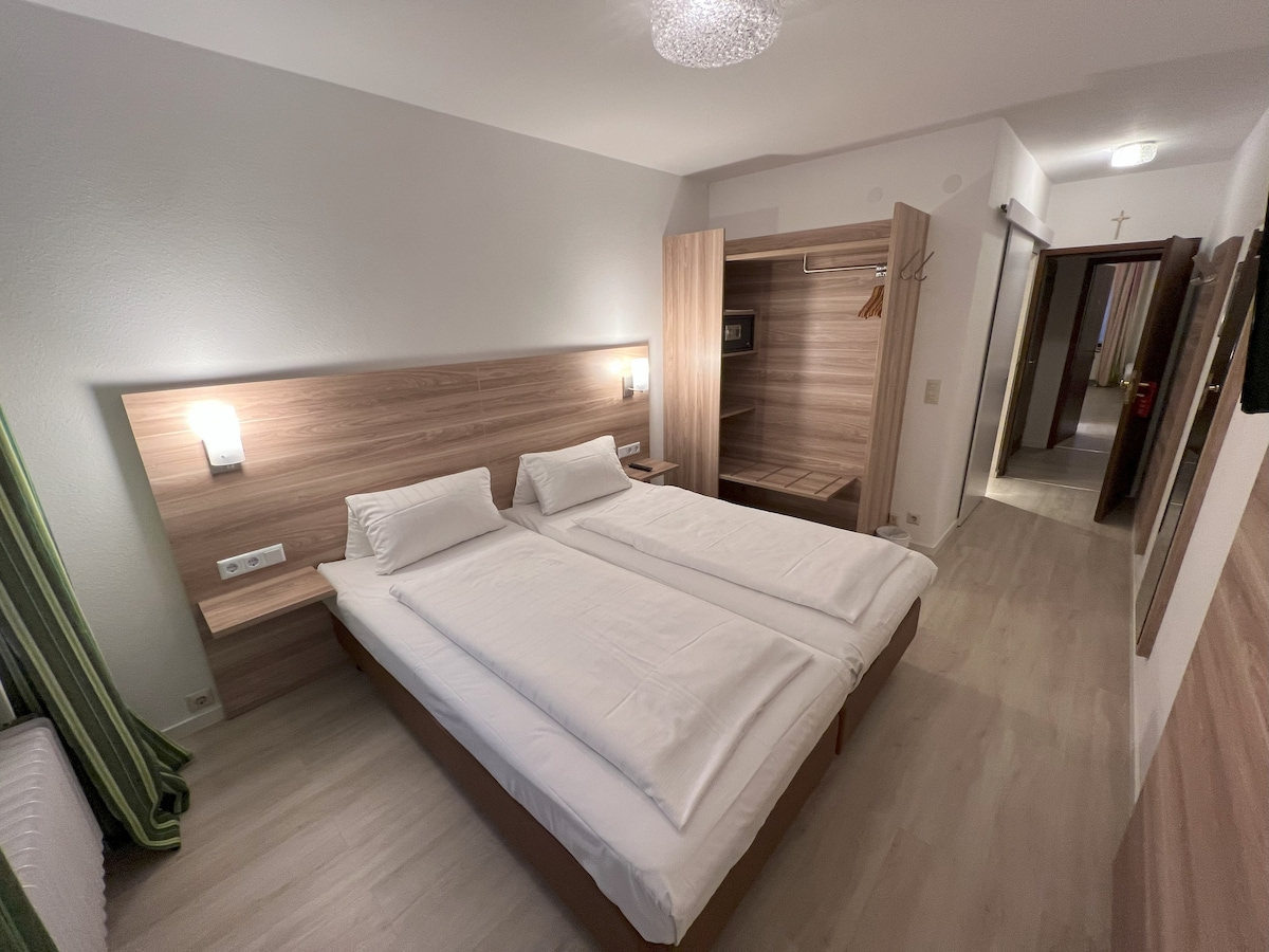 A well-lit sleeping area features two single beds with crisp white linens, positioned against a warm wood-panelled wall. A wardrobe and coat hook are visible in the background. The space is designed for comfort and functionality, with soft green curtains adding a touch of color.
