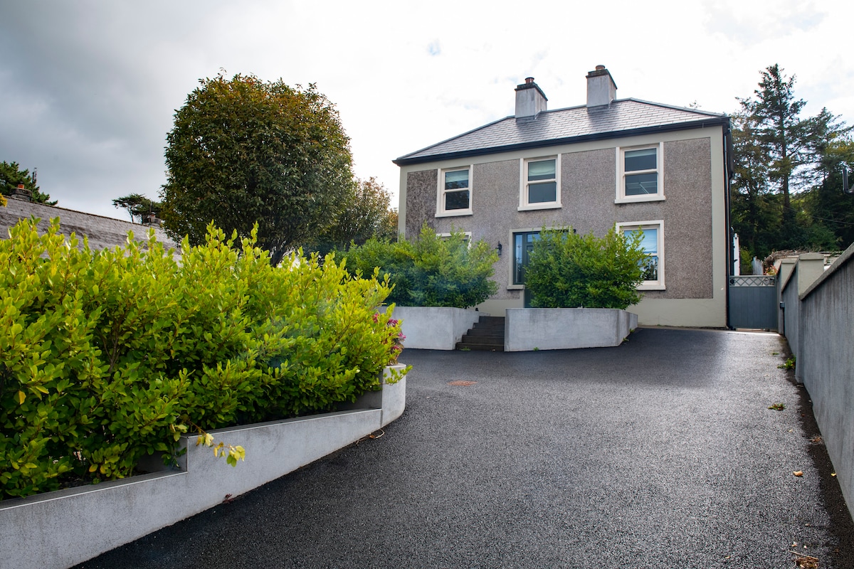 The exterior of a two-story house is framed by lush green shrubs and a well-maintained driveway. The pale gray façade features large windows, and the entrance is accentuated by a few steps leading up to the front door. A serene atmosphere surrounds the property.