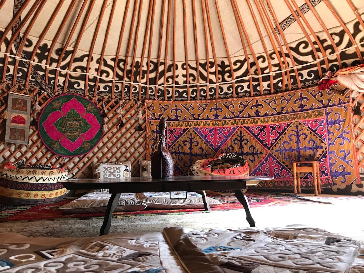 The interior of the yurt is adorned with vibrant wall decorations and traditional Kazakh patterns. A low wooden table occupies the center, surrounded by cozy woolen mats where guests can rest. Colorful textiles enhance the cultural atmosphere, creating a unique living space.