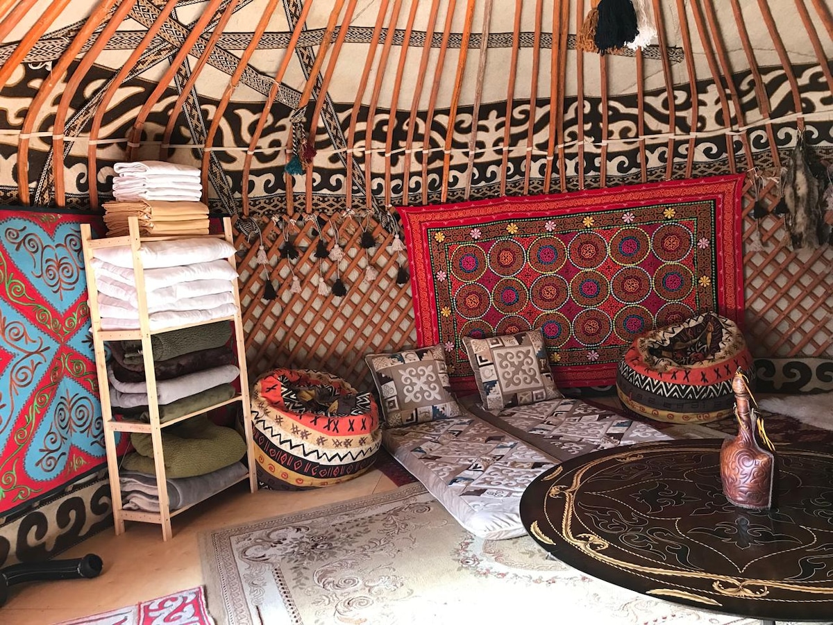 The interior of the yurt features intricate wall patterns and vibrant textiles. A round table sits at the center, surrounded by comfortable floor cushions. A neatly stacked shelf holds blankets and linens, while decorative items add cultural charm to the space.