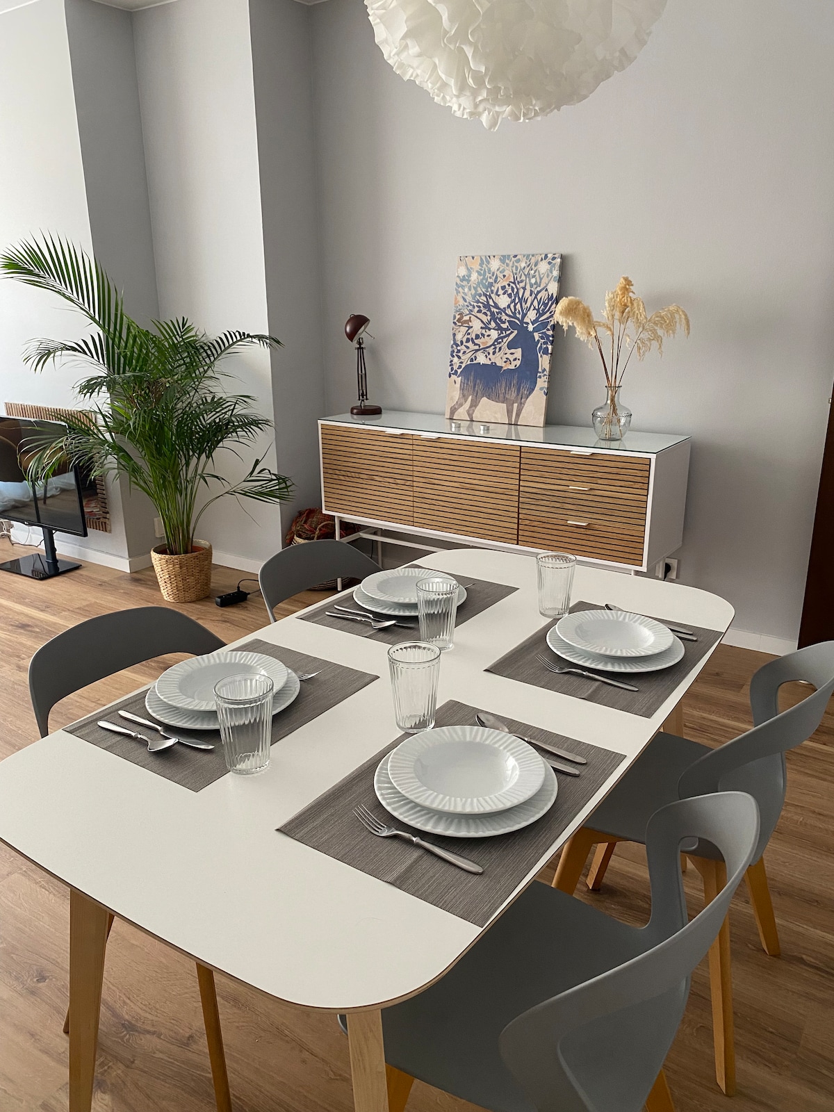 A dining area features a modern white table set for six, complete with plates, napkins, and glasses. A stylish sideboard displays decorative items, while a potted plant adds a touch of greenery. Natural light illuminates the space, enhancing its warm atmosphere.