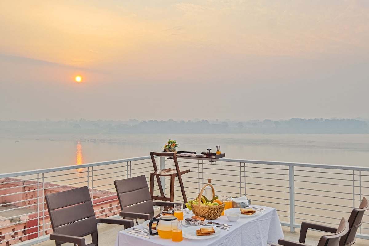 A spacious terrace is set for a meal with a table adorned with a white tablecloth, featuring breakfast items and beverages. The sun rises above the Ganges, casting a warm glow over the scenic landscape, enhancing the tranquil ambiance.