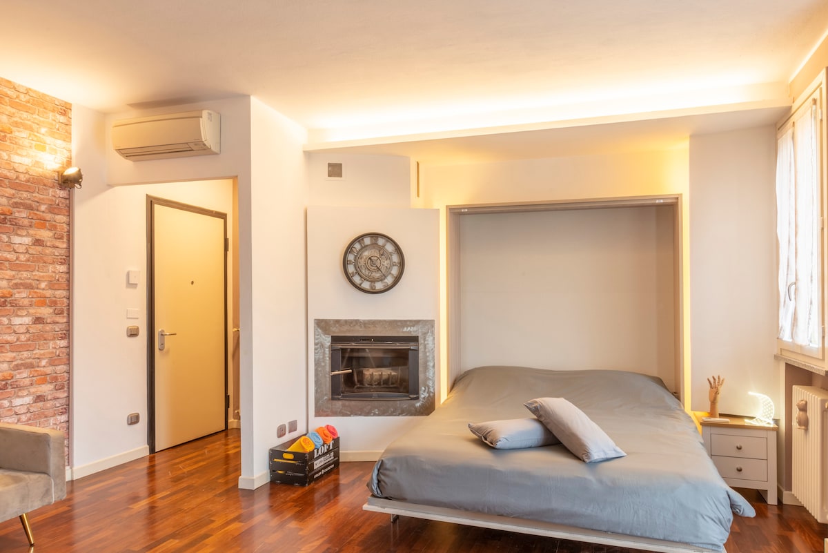 A spacious bedroom is featured, with a large bed covered in soft gray bedding. A decorative clock is mounted on the wall, and a fireplace is integrated into the design. Natural light enters through a window, enhancing the warm atmosphere of the room.