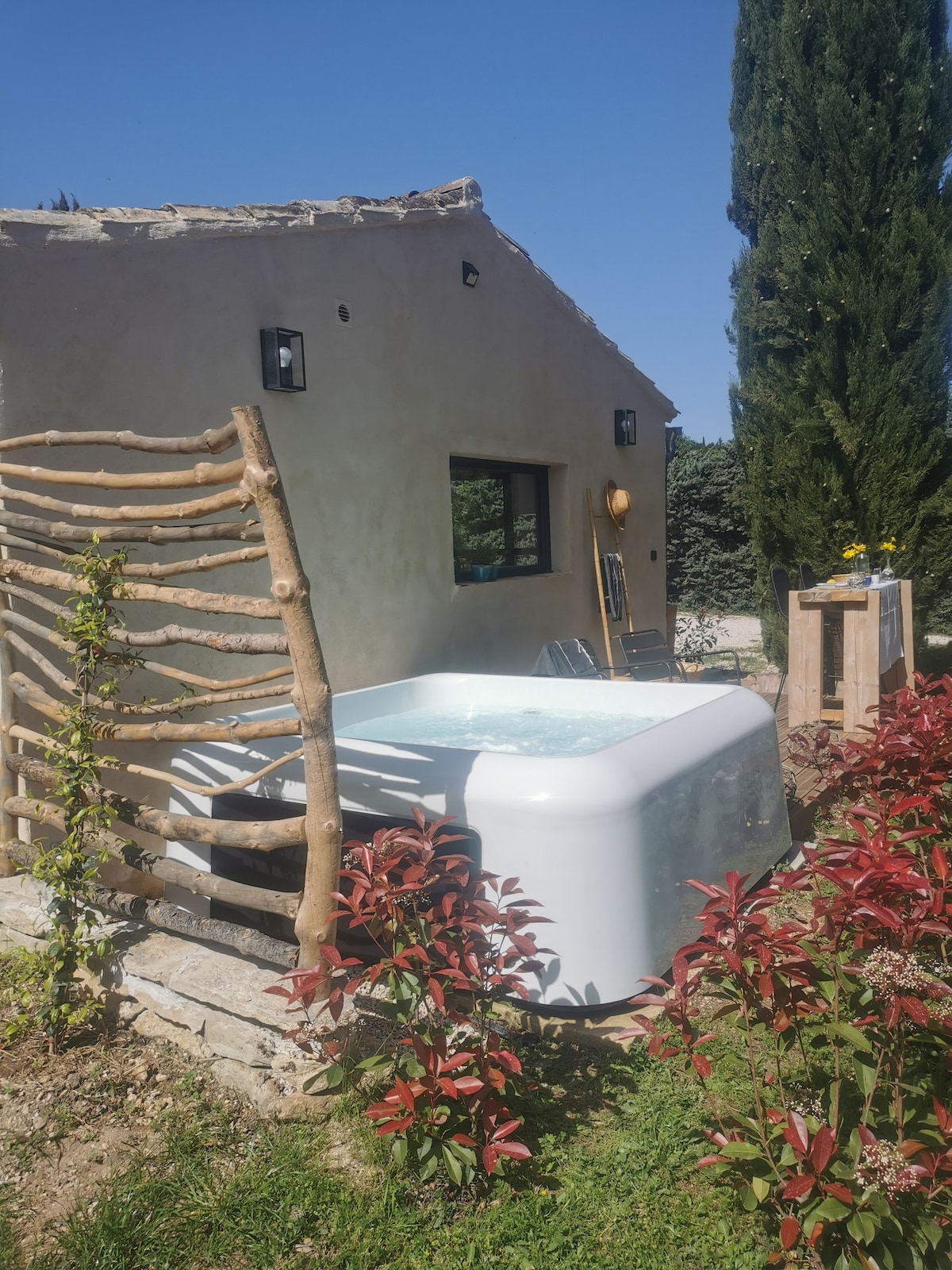 A private spa sits outside a charming structure, surrounded by lush greenery and colorful plants. Natural wooden elements frame the area, while sunlight highlights the tranquil setting. A sun chair is visible nearby, enhancing the inviting outdoor space for relaxation.
