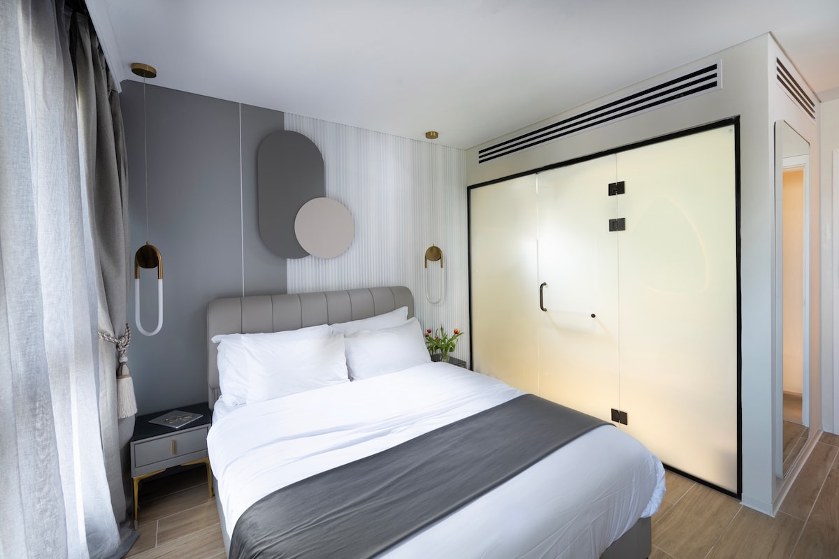 A modern bedroom features a spacious bed dressed in white linens with a contrasting gray throw. Soft curtains frame a window, while wall-mounted lights provide subtle illumination. A stylish, frosted glass sliding door separates the room from the adjoining space, adding a contemporary touch.