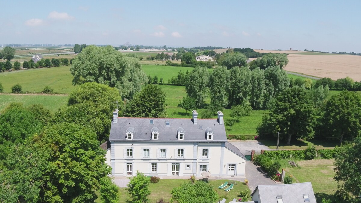An expansive property is showcased, surrounded by lush greenery and fields. The elegant white facade of the building stands out against a clear blue sky. Trees frame the landscape, creating a serene atmosphere, while the exterior hints at comfortable spaces for relaxation.