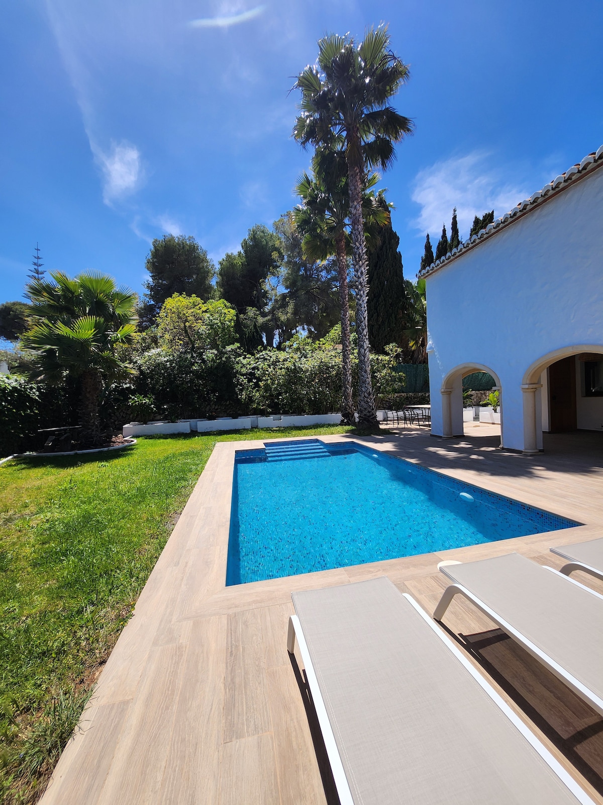 A private outdoor space features a clear blue swimming pool surrounded by lush greenery. Light-colored sun loungers are positioned near the pool, offering relaxation options. Tall palm trees and mature shrubs create a serene atmosphere, complemented by the architecture of the adjacent house.