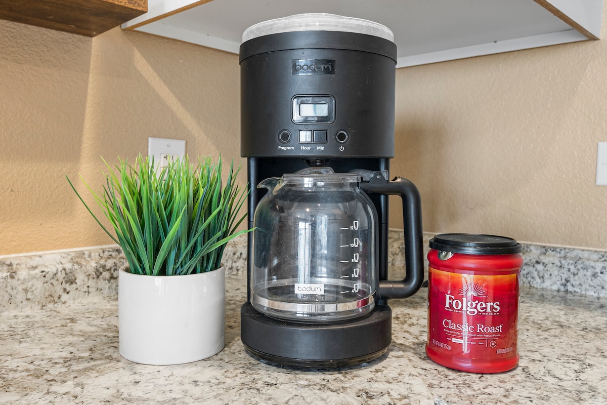 A coffee maker is positioned on a granite countertop, accompanied by a jar of Folgers Classic Roast coffee. A small potted plant adds a touch of greenery, enhancing the space's functionality and inviting appeal.