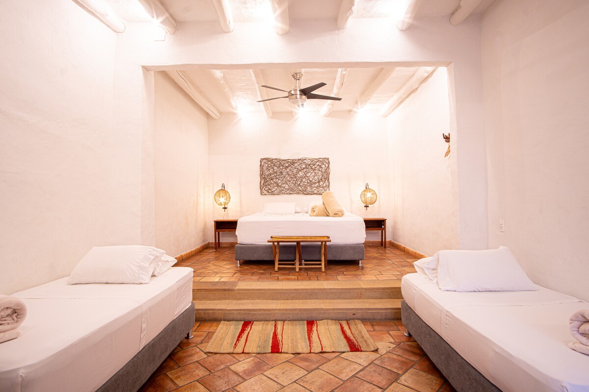 A spacious sleeping area features a large bed against a white wall, accompanied by two single beds on either side. A fan hangs from the ceiling, and warm light from decorative lanterns enhances the neutral decor. A patterned rug is laid across the tiled floor.