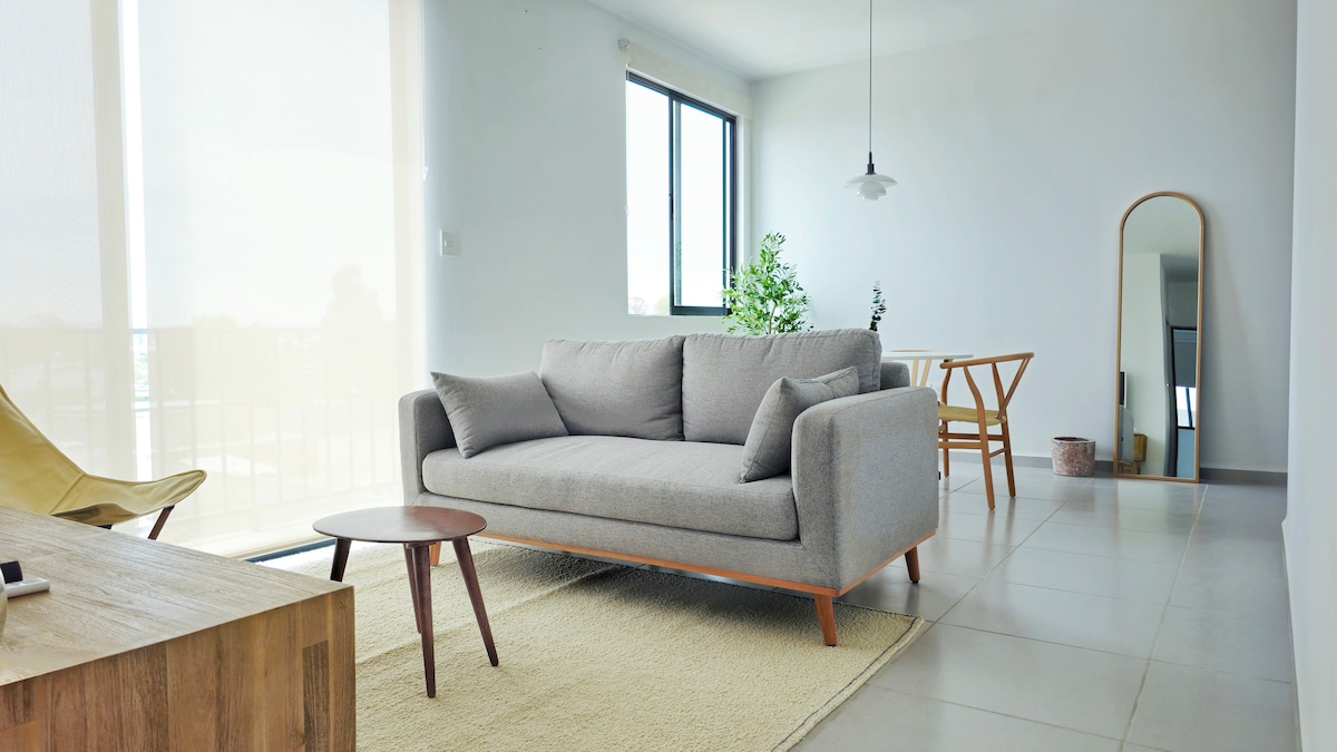 A light-filled living area features a gray sofa with two accent pillows, positioned on a neutral rug. A wooden coffee table and a dining area with a simple wooden table and chairs complement the minimalist design. Large windows allow natural light to brighten the space.