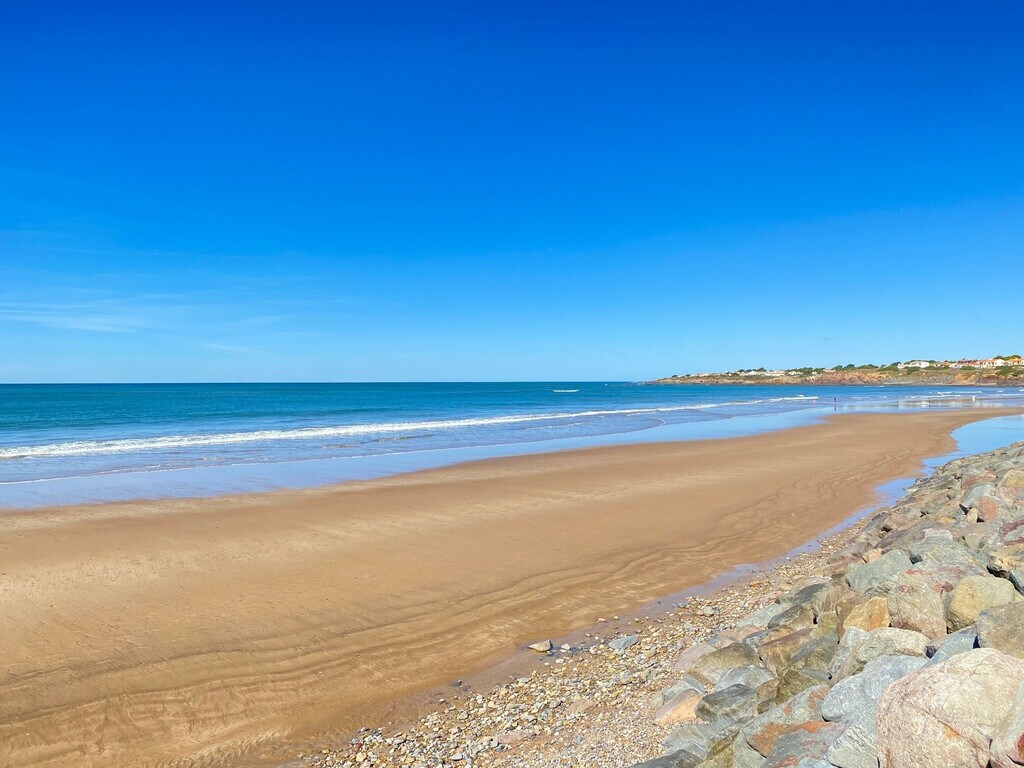 A wide beach is shown with smooth, golden sand stretching along the shore. Gentle waves lap at the water's edge, while a clear blue sky meets the horizon. The rocky coastline is visible in the foreground, providing a natural frame for the scenic view.