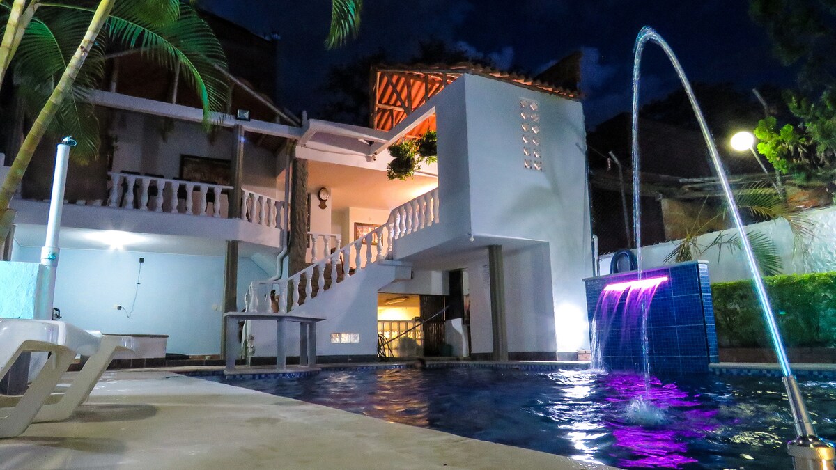 A modern building features a spacious pool illuminated with a soft purple light. Water cascades from a fountain, creating a calming ambiance. Lush greenery surrounds the area, complemented by decorative elements on the terrace above, enhancing the overall serene environment.