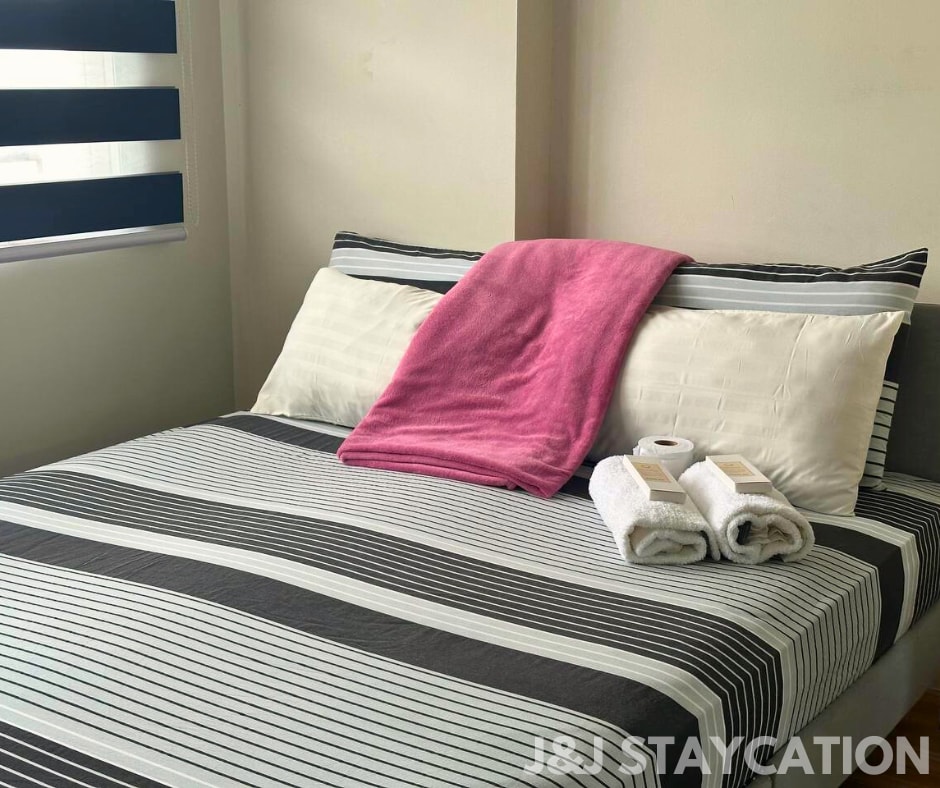 A spacious bed is dressed in striped bedding with neutral and bold tones. Two neatly arranged towels and toiletries are placed at the foot of the bed. A large window with dark roller blinds allows soft light to enter the room.