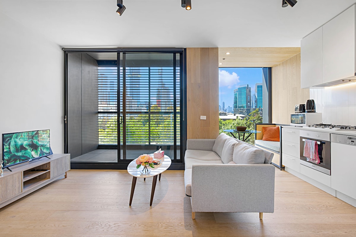 A modern living area is furnished with a light-colored sofa and a low coffee table complemented by a floral arrangement. Large glass sliding doors provide access to a balcony, allowing ample natural light and showcasing views of surrounding buildings and greenery.