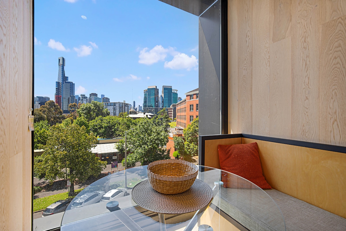 A dining area is positioned next to large windows, offering views of the city skyline and greenery outside. A round glass table is complemented by a woven basket and an orange cushion on a light wooden bench, enhancing the space's modern aesthetic.