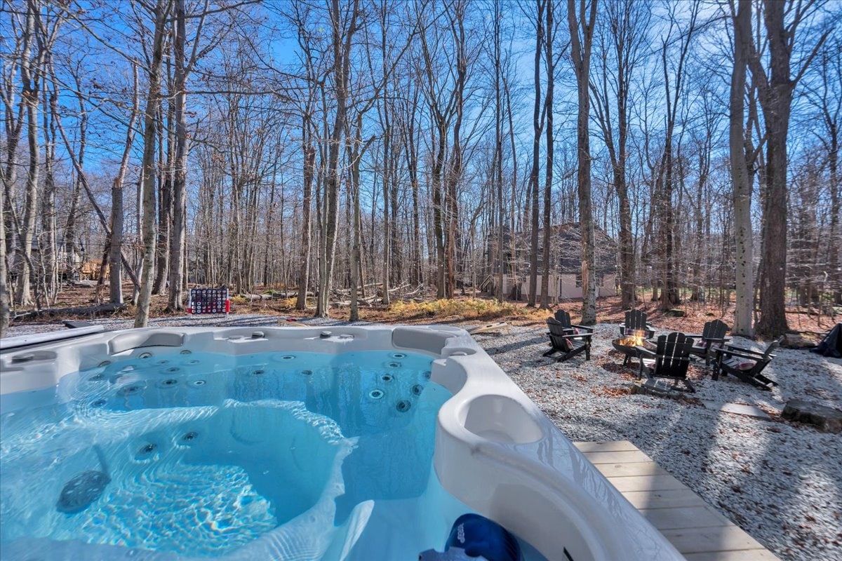 A hot tub is positioned on a wooden deck, surrounded by a wooded area with tall trees. An outdoor fire pit area is visible in the background, equipped with several Adirondack chairs, ideal for gatherings in a serene, natural setting.
