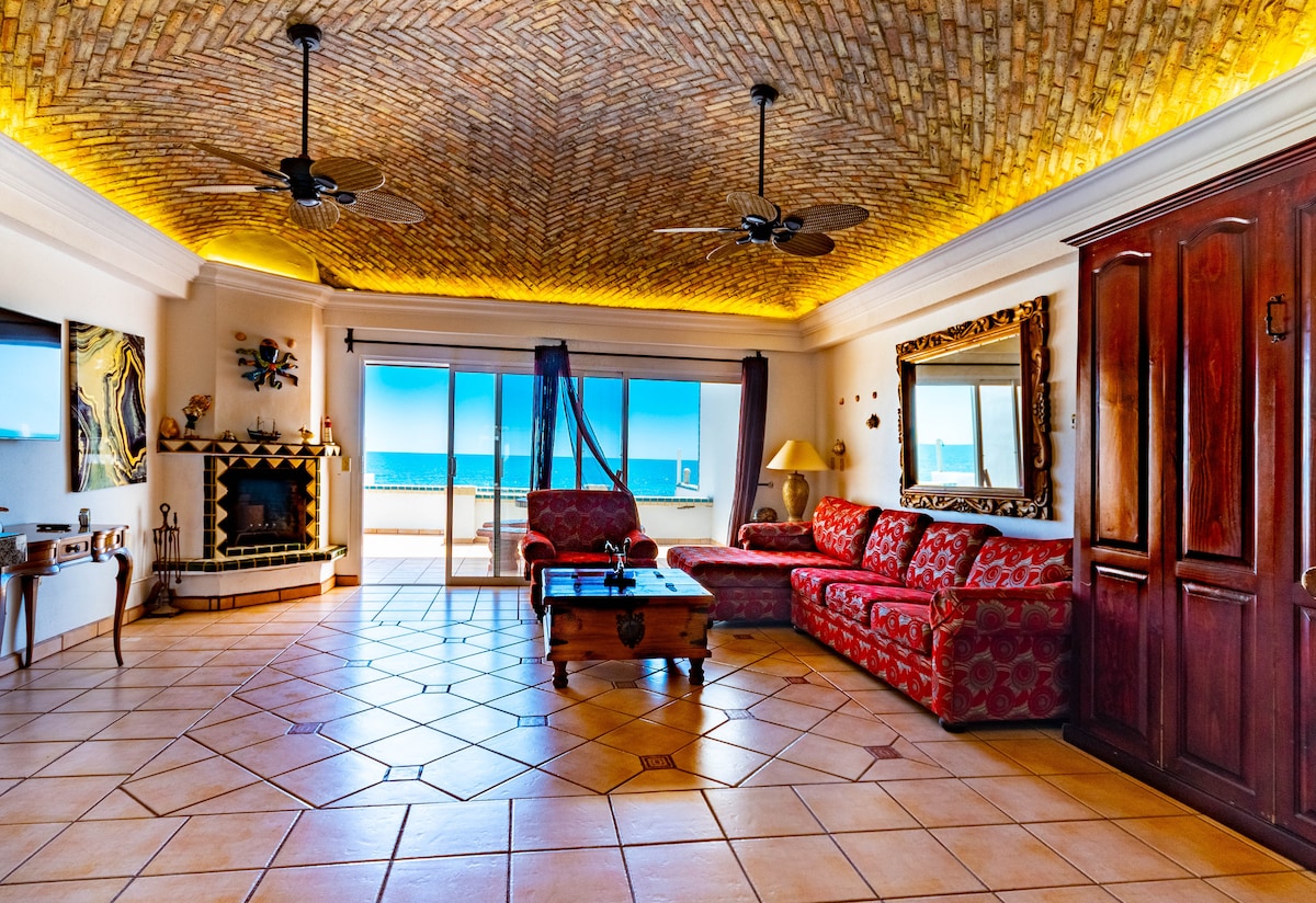 A spacious living area features a patterned ceiling with soft lighting and two ceiling fans. Red upholstered sofas are arranged around a wooden coffee table, complementing the tiled floor. Large glass doors offer views of the ocean, allowing natural light to fill the room.