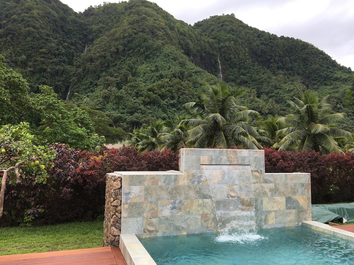 A tranquil swimming area is visible, designed with natural stone elements. It features a calm water feature cascading gently into the pool. The backdrop showcases lush green mountains, framed by vibrant tropical foliage and palm trees, creating a serene environment.