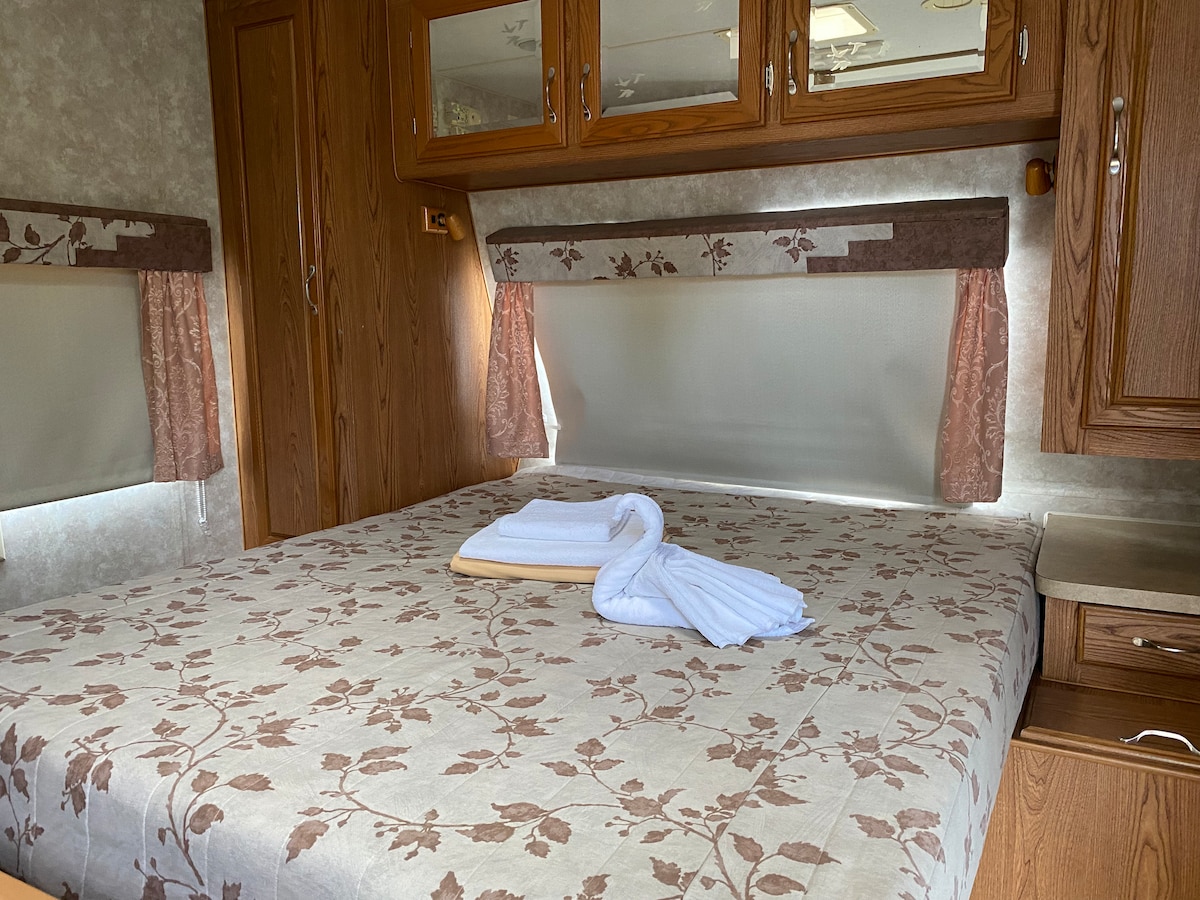 A spacious sleeping area is presented with a floral-patterned bedspread, complemented by neatly arranged towels. Wooden cabinetry and panels create a warm ambiance, while curtains filter light from the windows, allowing for a serene environment.