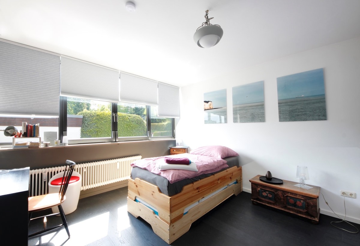 A bright bedroom features a wooden bed with a light pink comforter. Large windows with shades allow natural light to fill the space. A small bedside table and a chair are included, while artwork adorns the wall above the bed, enhancing the minimalist decor.