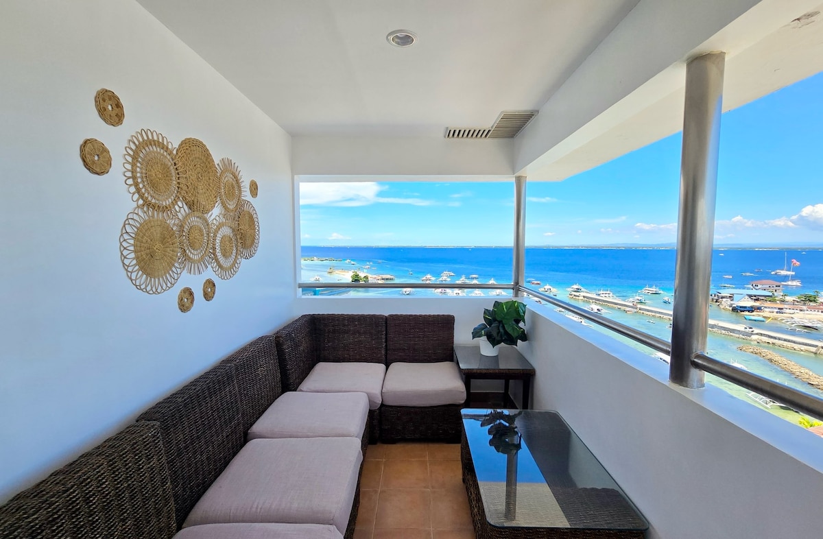 A seating area is situated on a balcony, featuring a brown wicker sofa with soft cushions and a glass-topped table. Views of the ocean and city landscape are visible through large windows, while decorative woven wall art adds a touch of local charm.