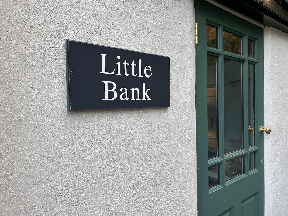 A sign reading 'Little Bank' is mounted on a wall beside a green door. The door features several panes of glass and a round gold doorknob, while the surrounding wall is finished in a smooth, light-colored texture.