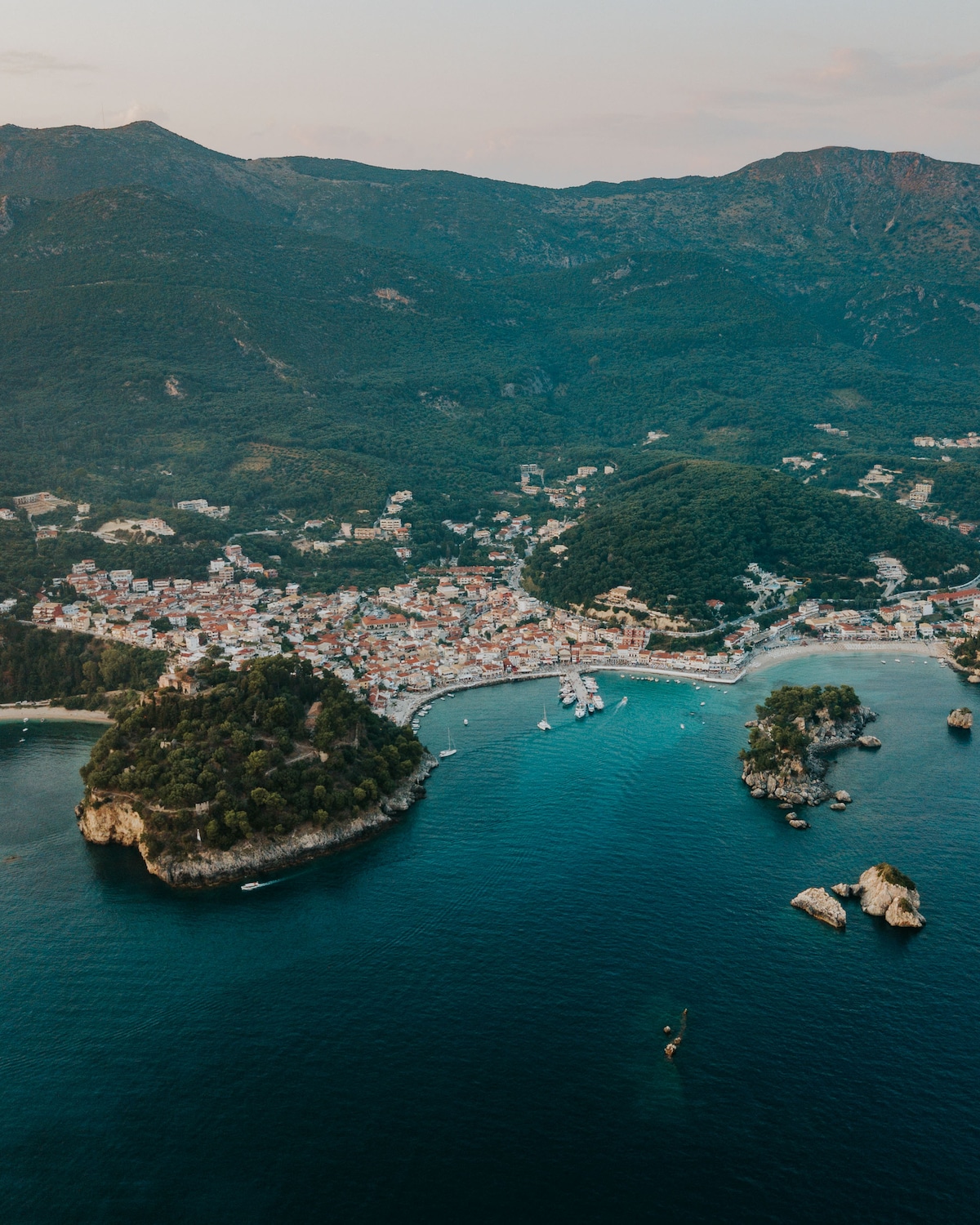 An aerial view captures the coastal town surrounded by lush green mountains. The winding coastline features a mix of sandy beaches and rocky formations, with boats dotting the tranquil turquoise waters. The layout of the town reveals clusters of buildings and roads leading to the harbor.