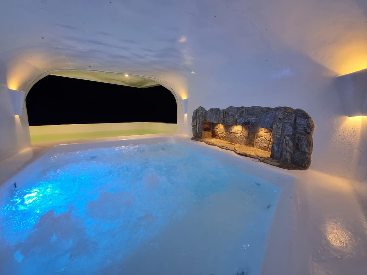 A grotto-style hot tub sits within a softly lit space, featuring an inviting hydrotherapy area. The walls are sculpted and illuminated with subtle lighting, while a stone feature runs along one side, enhancing the serene ambiance of the setting.