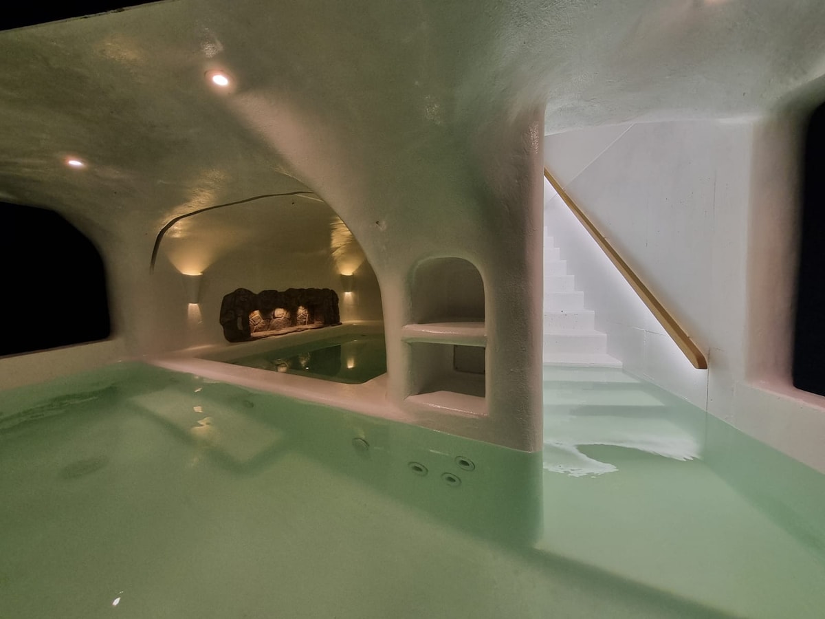 An inviting grotto features calming green water, soft illumination from recessed lights, and smooth, curved walls. A staircase leads upwards, while a stone feature offers a natural accent. This serene space provides a tranquil environment for relaxation and hydrotherapy.
