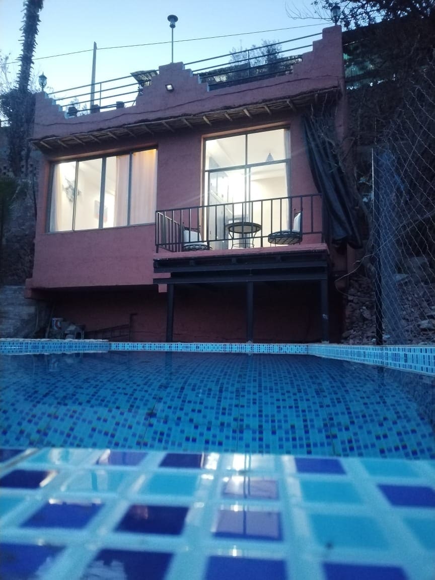 A charming structure is seen from the edge of a swimming pool, showcasing large glass windows that allow natural light to enter. A small balcony is present, featuring two chairs. The pool is framed by vibrant blue tiles, creating a serene atmosphere.